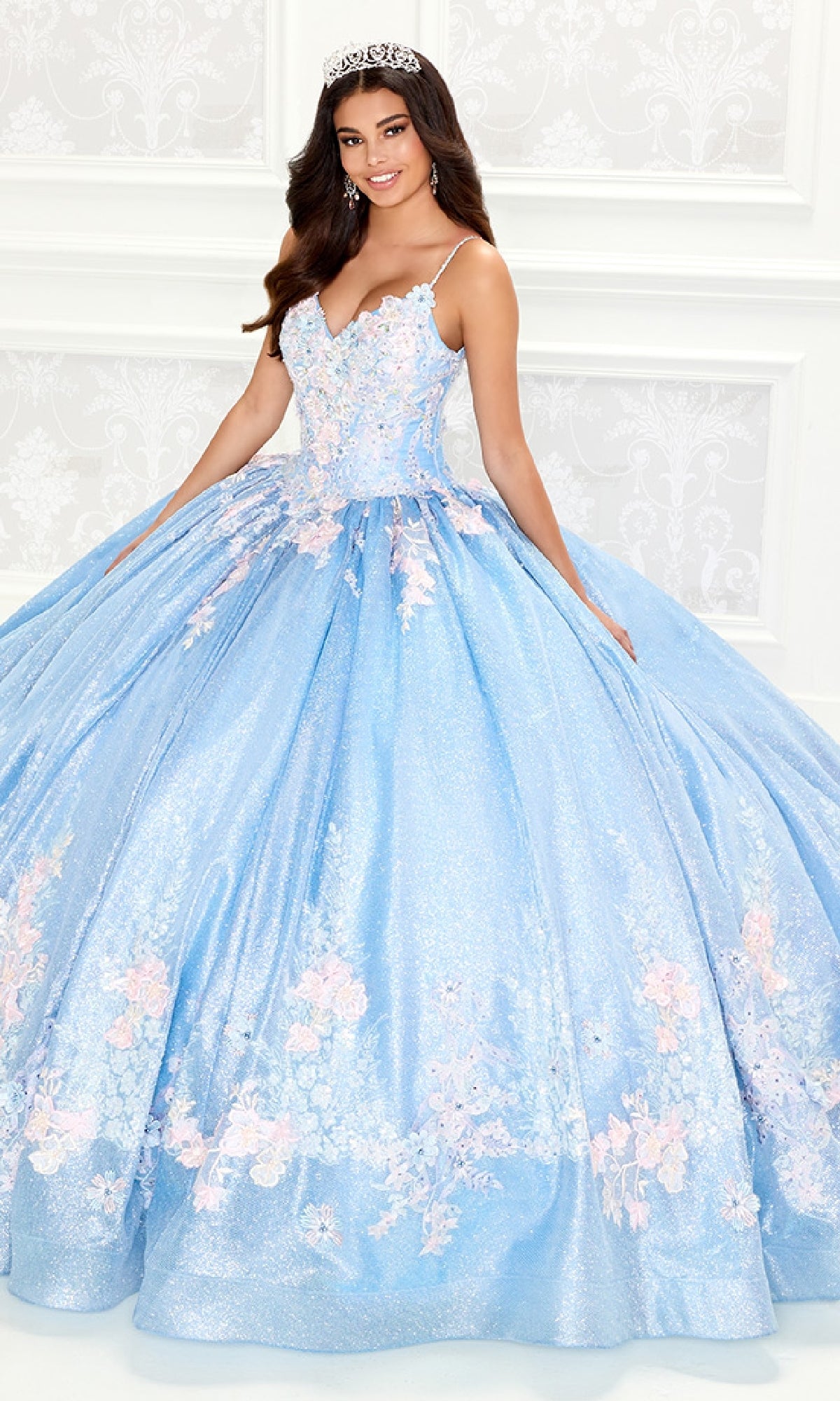 A young woman wearing the Light Blue Quinceanera Dress: Princesa PR30083, featuring a sparkling lace-embroidered corset bodice with floral details and a tiara, smiles as she stands before a white patterned wall.